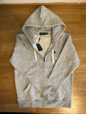 Grå hoodie från Polo Ralph Lauren - Snygg grå hoodie med dragkedja från Polo Ralph Lauren. Klassisk design med vit dragsko i huvan och mörkblå broderad logga på bröstet. Mjuk bomullsblandning och kängurufickor framtill. Perfekt för en chill och stilren look.