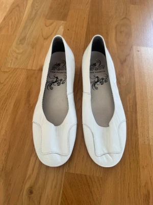 Vita ballerinaskor från Ten Points - Stilrena vita ballerinaskor från Ten Points i mjukt läder. Skorna har en fyrkantig tå och låg klack, samt diskreta sömdetaljer framtill. Insidan är dekorerad med ett svartvitt mönster och logga. Perfekta för en clean och klassisk look.