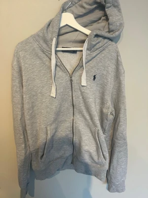 Grå hoodie från Polo Ralph Lauren - Snygg grå zip-hoodie från Polo Ralph Lauren med huva och dragsko. Klassisk logga broderad på bröstet och två fickor framtill. Tillverkad i mjukt bomullsmaterial som är perfekt för en avslappnad stil.