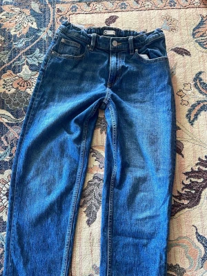Blå jeans med hög midja - Säljer ett par klassiska blå jeans med hög midja och raka ben. Jeansen har fem fickor och knappstängning framtill. Perfekt för dig som gillar en tidlös och enkel look. Materialet är denim och färgen är mörkblå.
