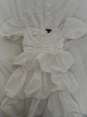 Vit volangklänning från Prettylittlething - Supersöt vit klänning från prettylittlething med puffärmar, volanger och markerad midja🌸i strl 36🌸jag har endast haft den på min student men köpte den här på plick🌸På sista bilden kan man se att själva ”underkjolen” är sönder på höger sida men det syns inte på något sätt när man har på sig den!🌸