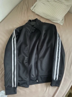 Träningströja - Svart träningsjacka från R med vita dubbla ränder längs ärmarna. Jackan har hög krage, dragkedja framtill och muddar vid ärmslut och nederkant. Perfekt för sport eller chill. Materialet är troligtvis polyester för en sportig känsla.