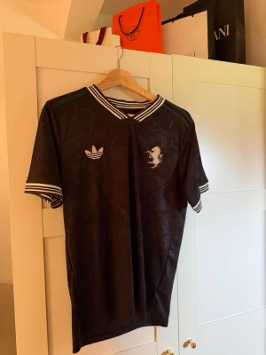 Svart Adidas t-shirt med mönster - Snygg svart t-shirt från Adidas med diskret blommönster i tyget. T-shirten har vita ränder på krage och ärmslut samt Adidas-logga och ett lejonmärke på bröstet. Perfekt för dig som gillar sportig och stilren design.