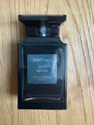 Tom Ford Oud Wood Eau de Parfum 100 ml - Exklusiv parfym från Tom Ford i doften Oud Wood. Flaskan är mörkgrå, nästan svart, med en elegant och stilren design i glas. Metallisk etikett framtill med märkets logga och doftnamn. Perfekt för dig som gillar lyxiga och unika dofter. Finns ca 80ml kvar därav rabatterad pris 