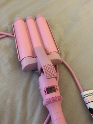Mermade Hair PRO Waver - Säljer en rosa vågtång med tre cylindrar och digital display. Tången har ergonomiskt handtag och knappar för temperaturkontroll. Perfekt för att skapa snygga beach waves. Tillverkad i plast och metall, med lång sladd för enkel användning.
