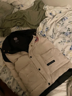 Beige dunväst från Canada Goose - Säljer en beige dunväst från Canada Goose med svart innerfoder och klassisk logga på bröstet. Västen har stora fickor framtill och stängs med både dragkedja och tryckknappar. Perfekt för dig som vill ha en stilren och varm väst till hösten eller vintern.