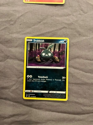 Pokémonkort Trubbish - Säljer ett Pokémonkort med Trubbish-motivet. Kortet har en färgstark design med svart, grönt och lila som dominerande färger. Det är tillverkat i laminerad papp och har en gul ram. Perfekt för dig som samlar eller spelar Pokémon TCG.