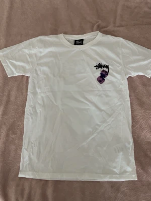 Vit Stüssy dice T-shirt med lila tärningar - Säljer en vit t-shirt från Stüssy med svart logga och lila tärningsmotiv på bröstet. Klassisk rund hals och korta ärmar. Perfekt för dig som gillar streetwear och vill ha något stilrent men ändå med en twist. 