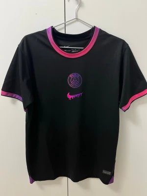Svart PSG x Nike t-shirt med rosa detaljer M - Snygg svart t-shirt från Nike i samarbete med PSG. Tröjan har rosa och lila detaljer vid krage och ärmslut, samt PSG-logga och Nike swoosh i rosa framtill. Tillverkad i lätt polyester som andas, perfekt för träning eller sport. Från CNFans 