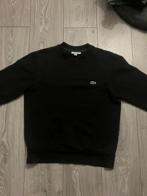 Svart sweatshirt från Lacoste - Klassisk svart sweatshirt från Lacoste med rund halsringning och den ikoniska gröna krokodilloggan broderad på bröstet. Pris kan sänkas via snabb affär.