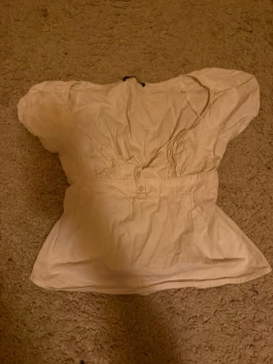 Vit blus med puffärm och v-ringning - Söt vit blus med puffiga korta ärmar och v-ringning. Blusen har knappar framtill och markerad midja som ger en feminin siluett. Tillverkad i ett lätt bomullsliknande material, perfekt för en trendig och avslappnad look.