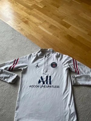 Vit PSG träningströja  - Säljer en vit långärmad träningströja från PSG och Jordan med dragkedja vid halsen. Tröjan har röda och blå ränder på ärmarna, PSG-logga på bröstet och tryck med 'Accor Live Limitless' samt 'Visit Rwanda' på ryggen. Perfekt för fotbollsträning eller chill.