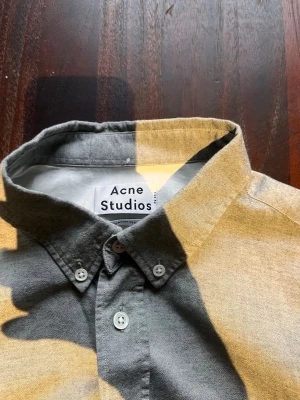 Grå skjorta från Acne Studios - Säljer en stilren grå skjorta från Acne Studios med klassisk krage och vita knappar. Skjortan har lång ärm och är tillverkad i ett mjukt bomullstyg. Perfekt för dig som gillar minimalistisk och clean stil.