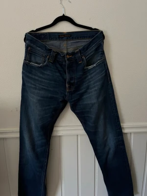 Blå jeans från Nudie Jeans - Hål i framfickorna. Nypris 1700kr. Säljer ett par klassiska blå jeans från Nudie Jeans med raka ben och snygga slitningar. Modellen är Lean Dean. Jeansen har fem fickor, orange sömmar och Nudie-logga på bakfickan. Tillverkade i denim med en cool tvättad look och detaljer som ger en avslappnad vibe.