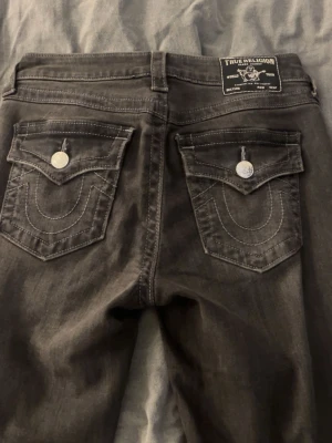 Svarta True Religion jeans med fickdetaljer - Snygga svarta jeans från True Religion med kontrastsömmar och stora ficklock bak med silverfärgade knappar. Modellen heter Becca och har klassisk femficksdesign, dragkedjegylf och bälteshällor. Perfekta för en cool och avslappnad stil. Är lite långa för mig som är 166