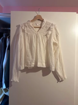 Vit blus med spets från Gina Tricot - Säljer en vit blus från Gina Tricot med romantiska detaljer som spets och rynkad krage. Blusen har långärmade ärmar och är tillverkad i ett lätt och luftigt material som känns skönt mot huden. Perfekt för dig som gillar en feminin och trendig stil.