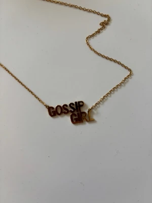Guldigt Gossip Girl-halsband - Säljer ett guldigt halsband från Rockin med texten 'GOSSIP GIRL' i metallbokstäver. Kedjan är tunn och elegant, perfekt för dig som gillar popkultur och statement-smycken. Ett riktigt coolt accessoar som sticker ut. Bara använd 1 gång. Pris går att diskutera!🥰