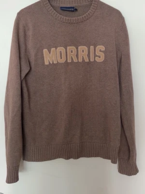 Beige tröja från Morris - Säljer en beige stickad tröja från Morris R. Club med stora bokstäver 'MORRIS' i ljusare beige på bröstet. Tröjan har rund halsringning och ribbade muddar vid ärmslut och nederkant. Perfekt för dig som gillar klassisk och enkel stil.