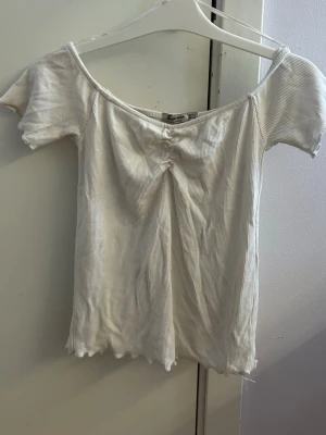 Vit ribbad topp med rynkdetalj - Säljer en vit ribbad topp från stradivarius med smala ränder och rynkdetalj vid halsen. Toppen har korta ärmar och en lätt offshoulder-design. Den är gjord i mjuk bomull och har vågig kant nertill för en extra touch.