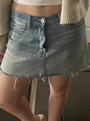 Blå jeanskjol med rå kant - Cool kort jeanskjol i ljusblå denim med rå, fransig kant och klassiska femficksdetaljer. Kjolen har knappstängning framtill och en avslappnad passform som är perfekt för en chill look. Passar dig som gillar en trendig och avslappnad stil.