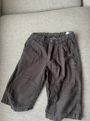 Bruna shorts från H&M med resårmidja - Säljer ett par bruna shorts från H&M med resår i midjan och knapp framtill. Shortsen har två sidofickor och är tillverkade i bomull, vilket gör dem sköna och lätta att bära. Perfekta för varma dagar och enkel att matcha med olika toppar.finns fler barnkläder på min sida
