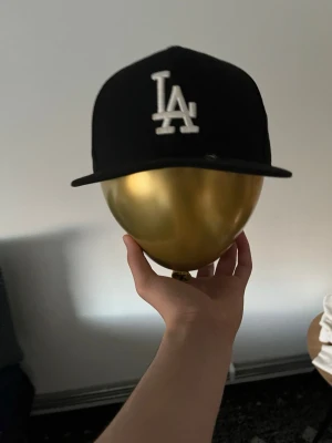 Svart LA 59FIFTY keps från New Era - Svart keps från New Era 59FIFTY med LA-logga broderad i vitt framtill. Klassisk flat brim och strukturerad passform. Tillverkad i slitstarkt tyg och har den ikoniska guldiga storleksstickern kvar på skärmen.