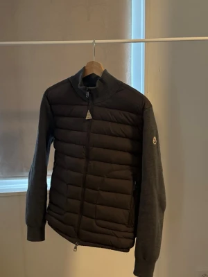Grå dunväst från Moncler - Snygg grå dunväst från Moncler med quiltad front och stickade ärmar. Västen har hög krage, dragkedja framtill och två sidofickor med dragkedja. Moncler-logga på ärmen ger en exklusiv touch. Perfekt för lager på lager och streetwear.