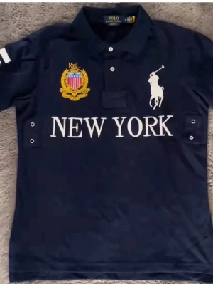 Mörkblå Polo Ralph Lauren skjorta - Mörkblå skjorta från Polo Ralph Lauren med klassisk krage, vita knappar och broderad logga på bröstet. Stora vita tryck med 'NEW YORK' och nummer 5 på ryggen. Skjortan har korta ärmar och detaljer i vitt och gult.