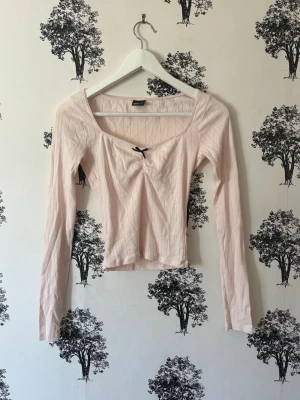Ljusrosa ribbad topp med rosett - Supersöt ljusrosa ribbad topp med lång ärm och liten svart rosett framtill. Toppen har en v-ringad urringning och croppad passform som ger en trendig look. Perfekt att styla med jeans eller kjol för en chill vibe.