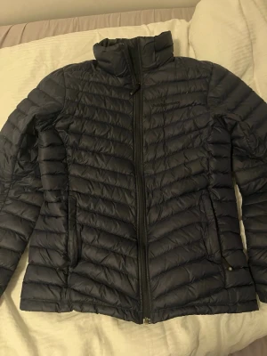 Svart dunjacka från Peak Performance - Snygg svart dunjacka från Peak Performance med diskret logga på bröstet. Jackan har hög krage, dragkedja framtill och två sidofickor med dragkedja. Lättviktigt material och quiltad design som håller värmen under kalla dagar.
