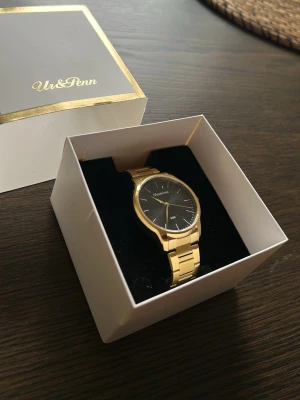 Guldfärgad klocka från Ur & Penn - Stilren klocka från Ur & Penn med guldfärgad metallarmband och svart urtavla. Klassisk rund form och minimalistisk design som passar till många olika stilar. Levereras i originalask.