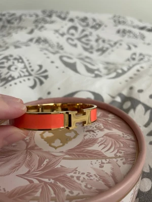  H armband - Armbandet är orange med guldfärgade metalldetaljer och har den ikoniska H-formade låset framtill. 