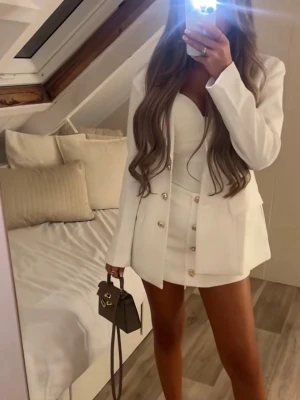 Vit kavaj och kjol med guldknappar - Säljer en stilren vit kostym med kavaj och matchande kjol. Kavajen har markerade axlar, klassisk krage och guldknappar framtill. Kjolen är kort och har samma snygga guldknappar. Perfekt för dig som vill ha en chic och modern look.