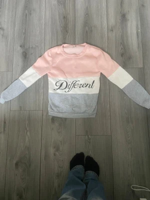 Rosa, vit och grå sweatshirt med text - Sweatshirt i färgblock med rosa, vitt och grått. Framsidan har texten 'Different' i svart skrivstil. Tröjan har rund halsringning och långa ärmar. Perfekt för en avslappnad och trendig look.
