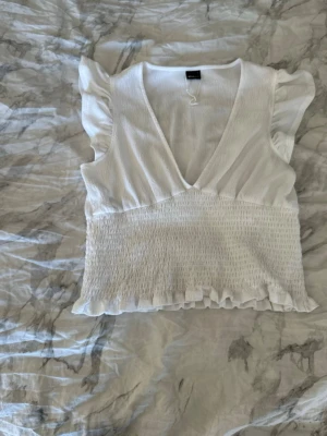 Vit blus med volang och v-ringning - Söt vit blus med djup v-ringning, volangdetaljer vid axlar och nederkant samt smockad midja för en feminin siluett. Ärmlös modell i lätt och luftig bomullsmix, perfekt för varma dagar. Enkel att styla till både jeans och kjol.