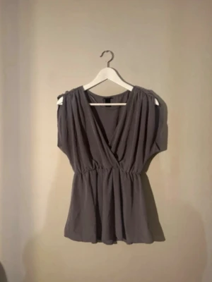 Grå omlottopp med v-ringning - Snygg grå omlottopp med v-ringad hals. Toppen är ärmlös och har ett luftigt, avslappnat fall. Perfekt att styla med jeans eller kjol för en trendig look.