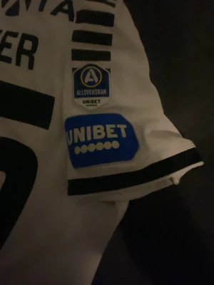 Vit fotbollströja Allsvenskan Sveijer match använd  - Säljer en vit fotbollströja med svarta detaljer och tryck, nummer 35 och namnet Sveijer på ryggen. Tröjan har Allsvenskan- och Unibet-loggor på ärmen samt flera sponsorer på ryggen. Perfekt för dig som gillar svensk fotboll och vill ha en snygg supportertröja.