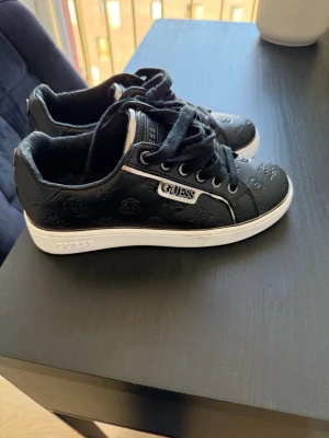 Guess sneakers  - Snygga svarta sneakers från Guess med präglad logga över hela skon och vita sulor. Skorna har svarta skosnören, metallfärgade detaljer och Guess-märke på sidan. Tillverkade i konstläder med en klassisk låg siluett.