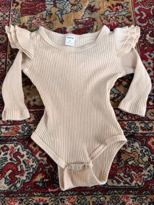 Beige ribbad body med volang SHEIN - Supersöt beige ribbad body från SHEIN med långa ärmar och volangdetaljer på axlarna. Tryckknappar i grenen gör den enkel att ta på och av. Perfekt för en trendig och bekväm look. Materialet är mjukt och stretchigt för extra komfort.