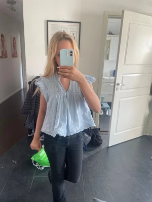 Ljusblå volangblus med v-ringning - Söt ljusblå blus med volangdetaljer vid ärmarna och v-ringad hals. Luftig och flowy passform, perfekt för varmare dagar. Tillverkad i ett tunt, mjukt bomullsliknande material. Passar dig som gillar en avslappnad men ändå söt stil.