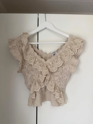 Beige topp från Zara - Supersöt populär beige topp från Zara med breda volangdetaljer och spetsmönster. Toppen är stickad/virkad och har en omlottkänsla framtill samt bred v-ringning. Liten i storleken, kom med prisförslag. 