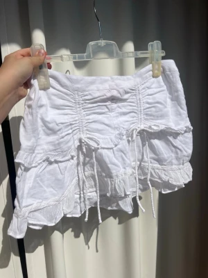 Vit volangkjol med snörning - Supersöt vit kortkjol med volanger och rosetter. Kjolen är i linnetyg så söt till sommar och midsommar 😍 använd typ 1 gång. Är från Ginas barn sektion är storlek 14+ skulle säga att det motsvarar L