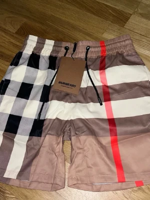Rutig shorts från Burberry - Säljer ett par snygga shorts från Burberry med klassiskt rutigt mönster i beige, svart, vitt och rött. Shortsen har resår i midjan med snörning och är tillverkade i ett lätt och bekvämt material. Perfekta för en trendig och avslappnad stil.
