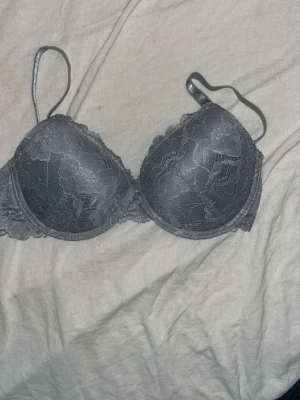 Grå spets-bh med vaddering - Snygg grå bh med vadderade kupor och spetsdetaljer över hela framsidan. Justerbara axelband och hakknäppning bak. Bh:n har en feminin design med mjuk spets och slät insida för extra komfort. 75B