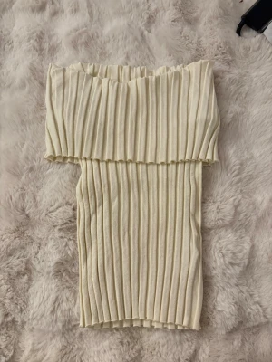 Offshoulder ribbad topp i beige - offshoulder topp från NA-KD i beige färg med ribbad struktur. Toppen är axelbandslös och har en croppad passform som sitter snyggt på kroppen. Perfekt för dig som vill ha en stilren och modern look. Storlek S men otroligt stretchig. Använd fåtal gåmger