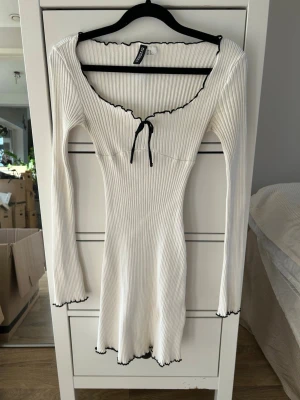 Vit ribbad klänning med svarta kanter och rosett - Supersöt vit ribbad kortklänning från HM med långa ärmar och svarta vågiga kanter. Klänningen har en djup v-ringning med knyt framtill och sitter figurnära.💗