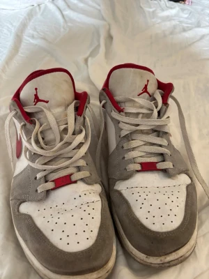 Nike Air Jordan 1 Low röd/vit/grå - Säljer ett par Nike Air Jordan 1 Low sneakers i vitt, grått och rött. Skorna har klassisk låg siluett, röd swoosh och Jumpman-logga på plösen, låda medföljer ej. Ovandelen är i mocka och läder med perforerad tåbox för ventilation. Perfekta för dig som gillar streetwear och snygg design.