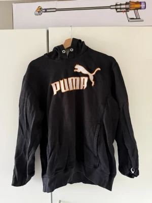 Svart hoodie från Puma med logga - Svart hoodie från Puma med stor vit och orange broderad logga på bröstet. Klassisk huva med dragsko och känguruficka framtill. Tillverkad i mjukt bomullsmaterial, perfekt för en avslappnad och sportig stil. Bra skick. Storlek L