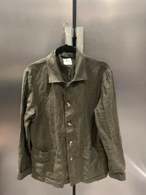 Olivgrön overshirt i linne från John Henric  - Stilren olivgrön overshirt från John Henric i linne. Klassisk krage, fyra knappar framtill och två stora fickor nedtill. Lätt och luftig modell med avslappnad passform, perfekt för lager på lager. Enkel design som funkar till många olika looks. Perfekt till sommaren eller våren!