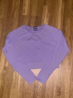 Ljuslila croppad topp från Aim'n - Säljer en ljuslila croppad topp från Aim'n med lång ärm och ribbad struktur. Toppen har en stilren look med rund halsringning och en liten metallbricka med logga på sidan. Perfekt för dig som gillar sportig och trendig stil.